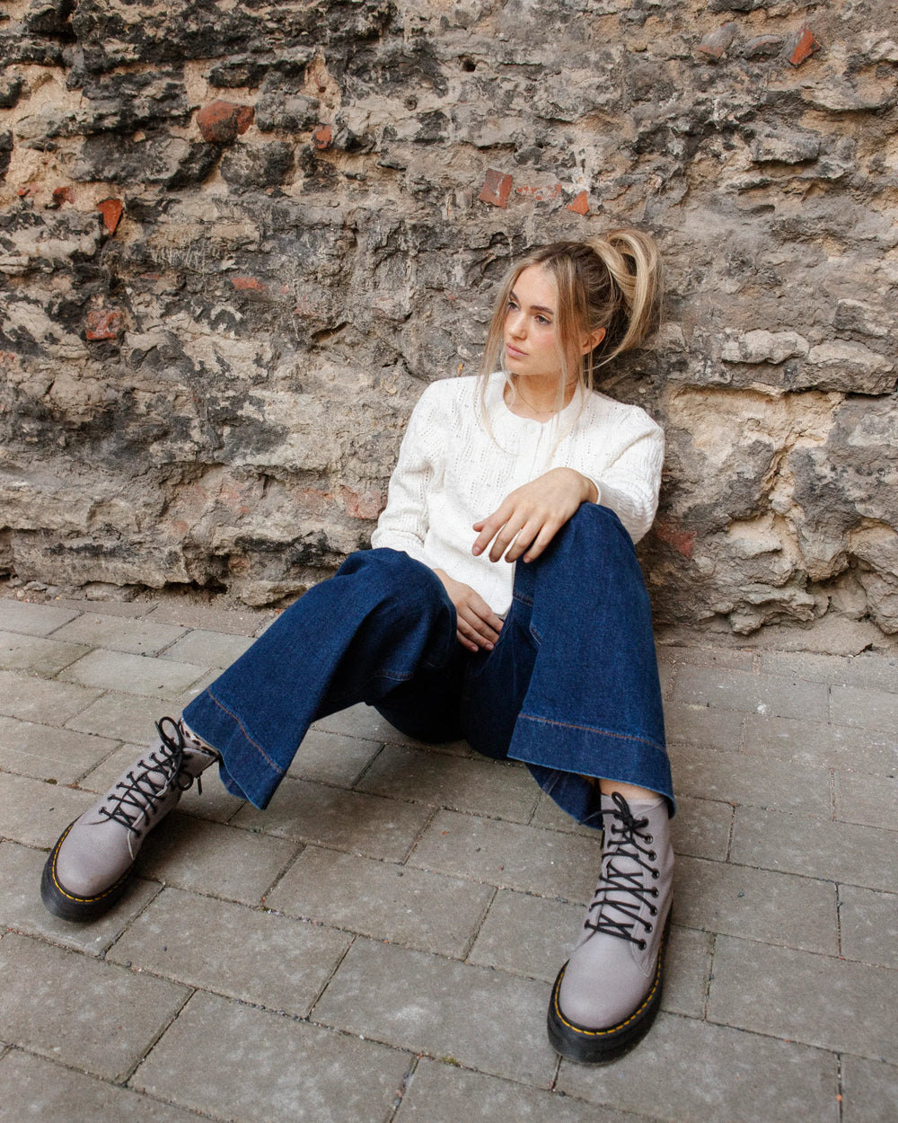 Full-length view of a woman seated against a stone wall wearing the Adele & Soleil Brise chalk white textured cardigan with dark trousers.