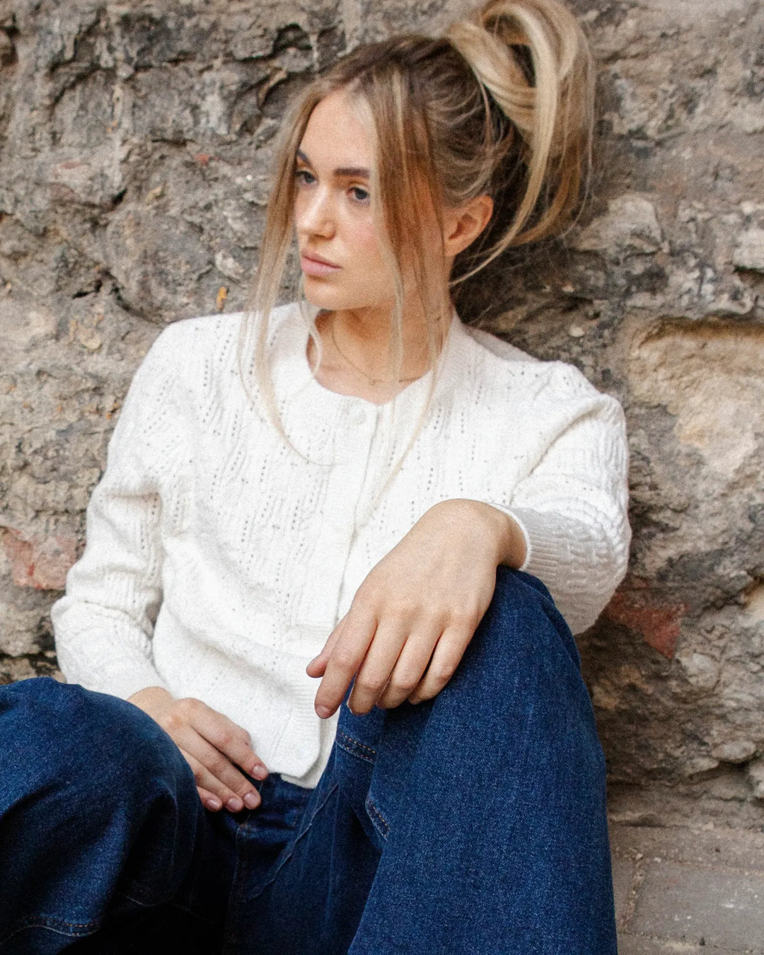 Woman seated against a stone wall wearing the Adele & Soleil Brise chalk white cardigan with a softly fitted silhouette.